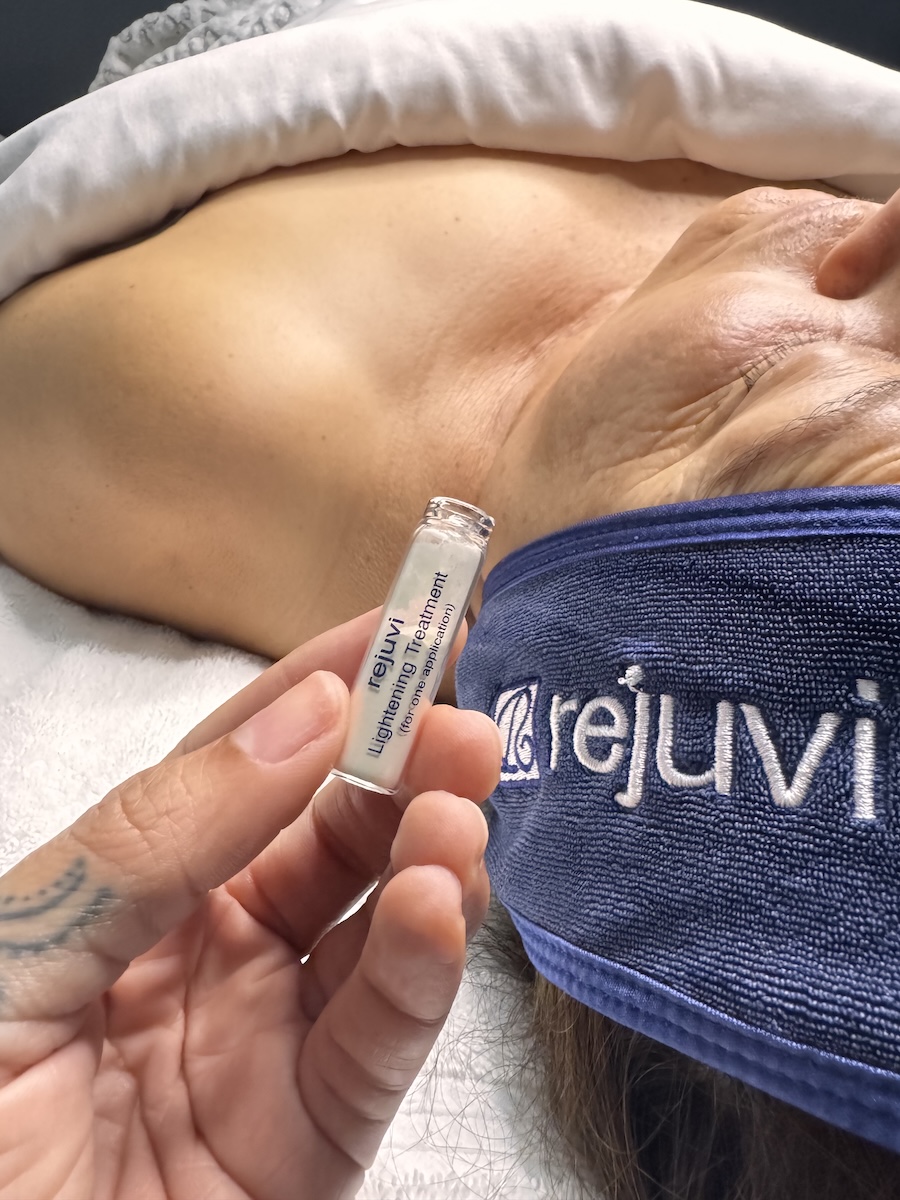 Hand holding up a product called rejuci lightening treatment during a facial with a client laying on the bed.